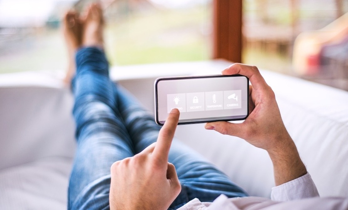 Eine Person liegt auf einem Sofa und steuert mit einem Smartphone Smart-Home-Funktionen. Auf dem Bildschirm werden Symbole für Funktionen wie Sicherheit und Beleuchtung angezeigt. Die Person trägt Jeans und ein langärmeliges Hemd, hat die Beine übereinandergeschlagen und ruhen auf dem Sofa.