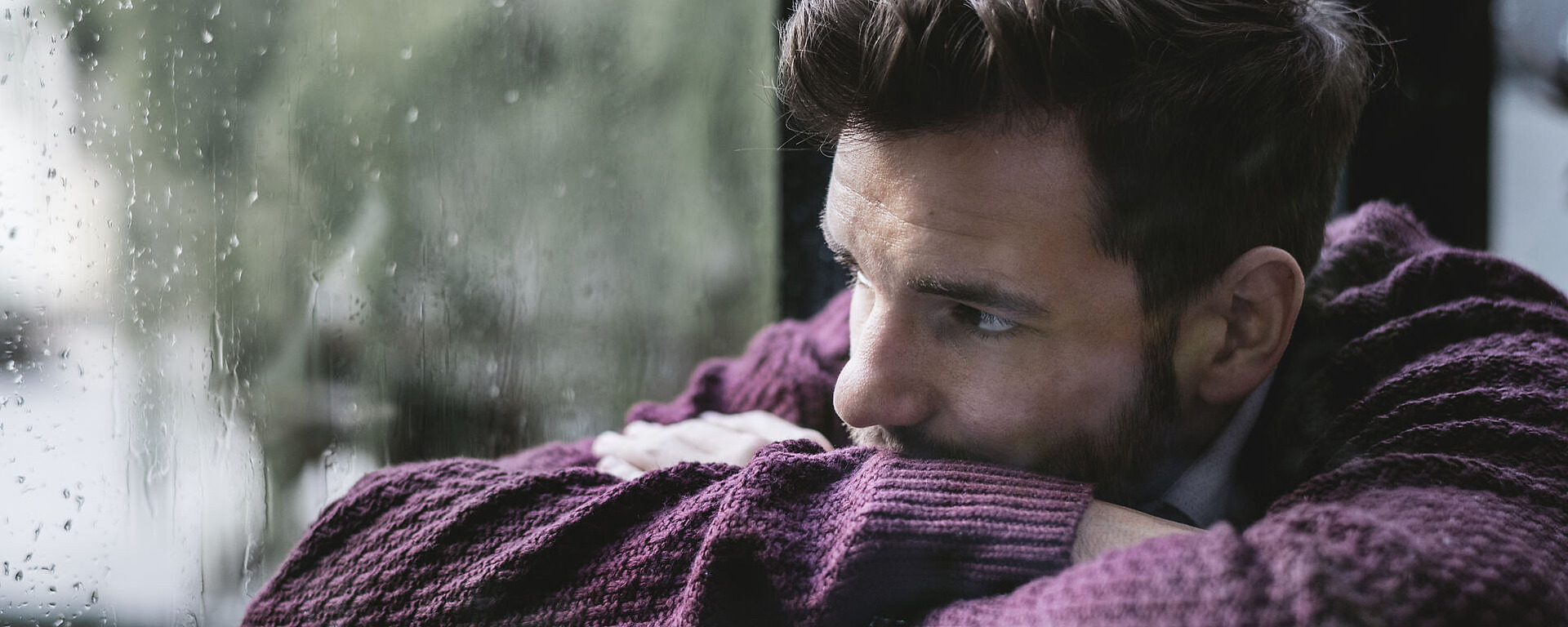 Ein Mann in einem kastanienbraunen Pullover sitzt in einem Haus, stützt die Arme auf die Knie und blickt nachdenklich aus einem regengesprenkelten Fenster. Die Stimmung ist kontemplativ und düster.