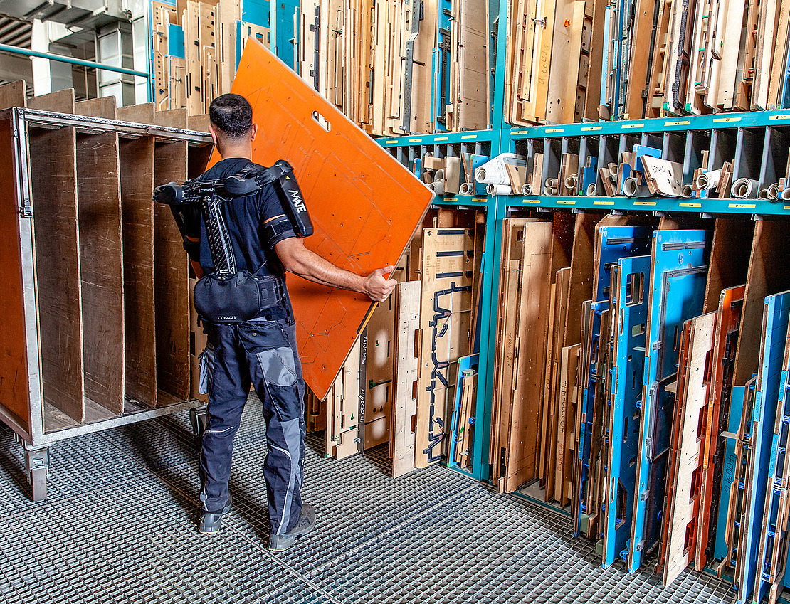 Eine Person, die einen schwarzen Exoskelett-Anzug trägt, hebt eine große orangefarbene Tafel in einem Lagerbereich an, der mit verschiedenen aufrecht stehenden Platten und Tafeln auf blauen Metallregalen gefüllt ist.