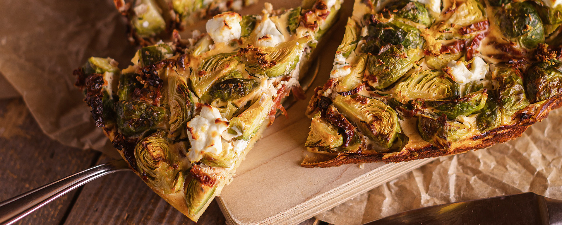 Zwei Scheiben herzhafte Torte mit Rosenkohl und zerbröckeltem Käse auf einem Holzbrett, daneben Gabel, Messer und loser Rosenkohl auf einem rustikalen Holztisch.