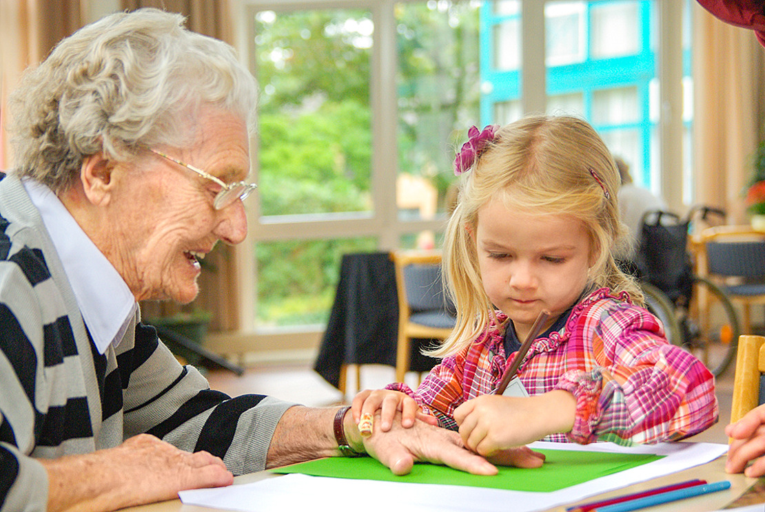 Eine ältere Frau und ein junges Mädchen sitzen an einem Tisch. Das Mädchen malt auf grünem Papier. Die Frau lächelt und schaut zu. Sie befinden sich in einem hellen Raum mit großen Fenstern. Draußen ist ein Garten zu sehen.