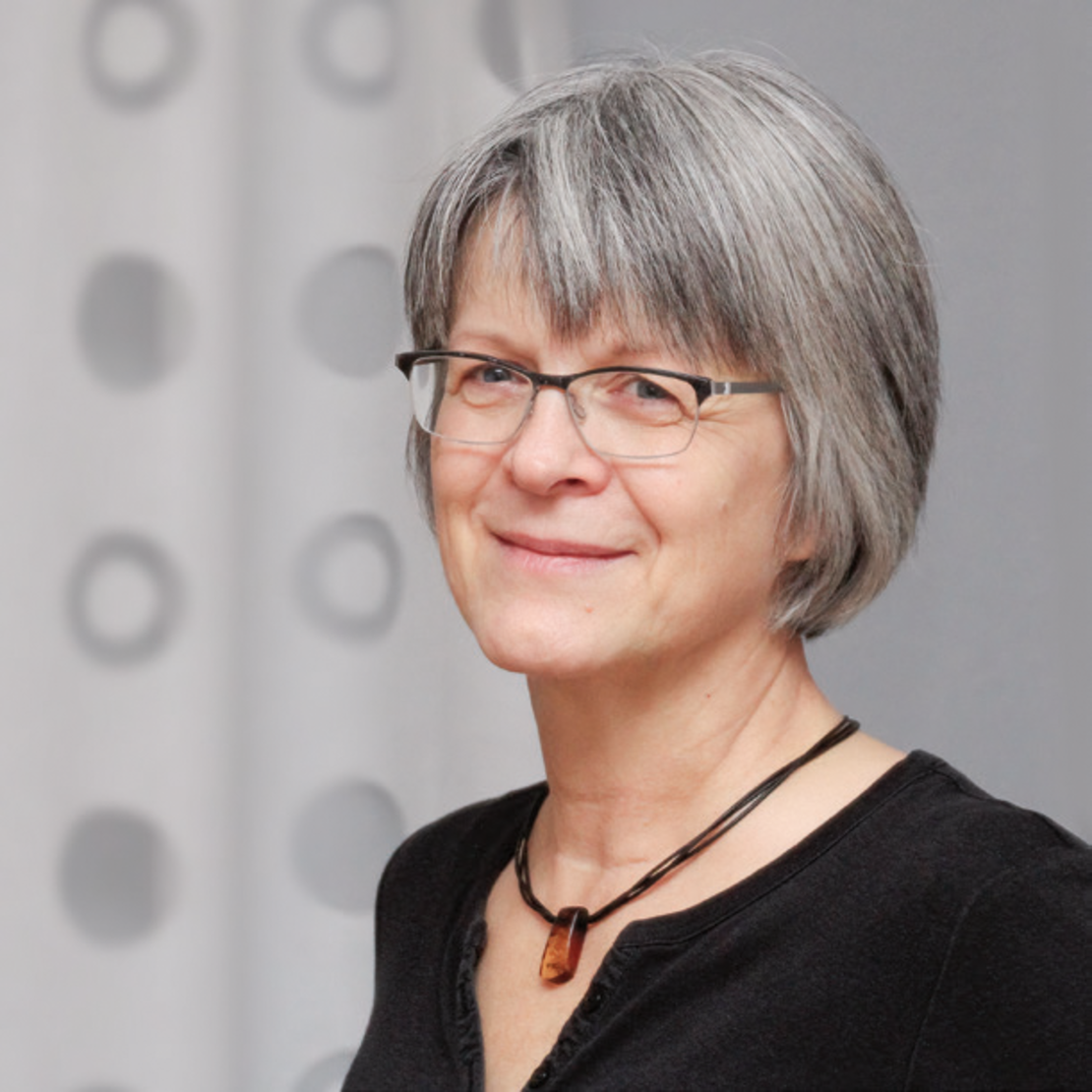 Eine lächelnde Frau mit kurzen grauen Haaren und Brille, die ein schwarzes Top und eine Halskette trägt, steht vor einem hellen Hintergrund mit kreisförmigen Mustern.