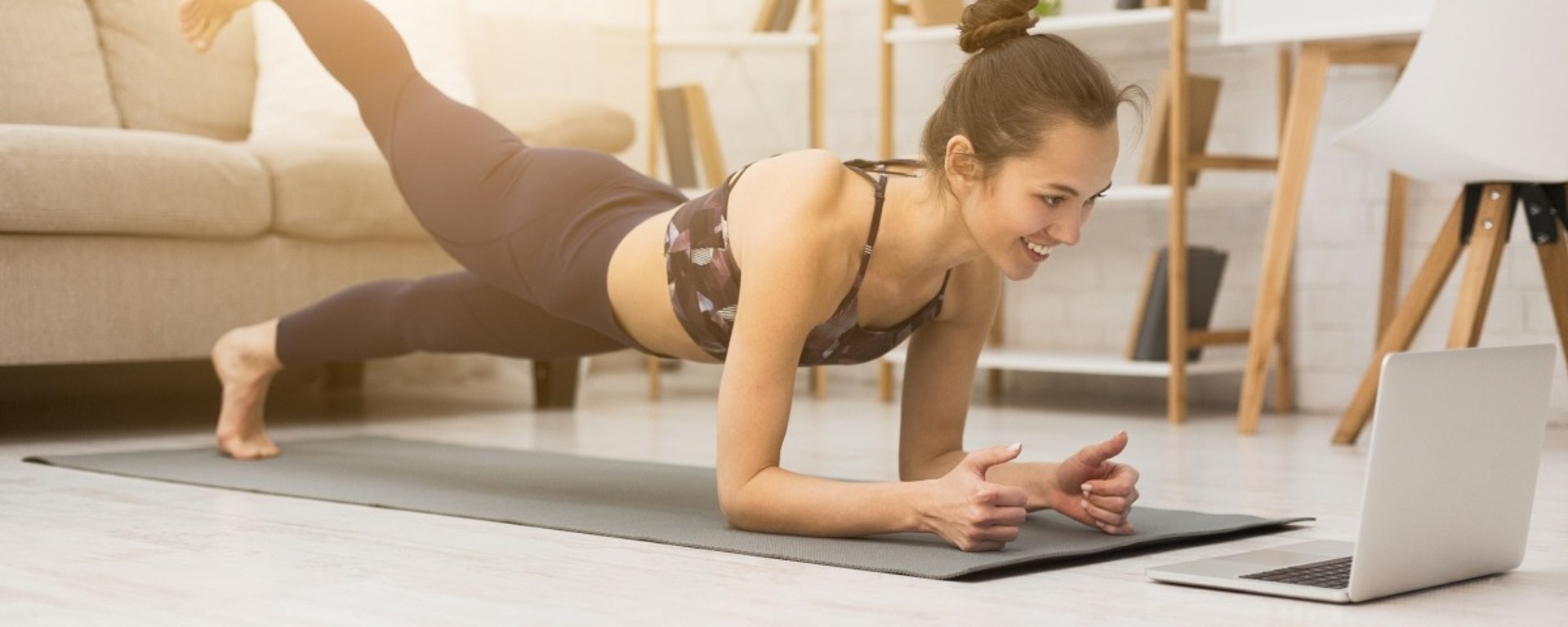 Eine Frau, die auf einer Yogamatte in ihrem Wohnzimmer eine Planke mit einem angehobenen Bein macht, während sie lächelt und auf den Laptop vor ihr schaut.