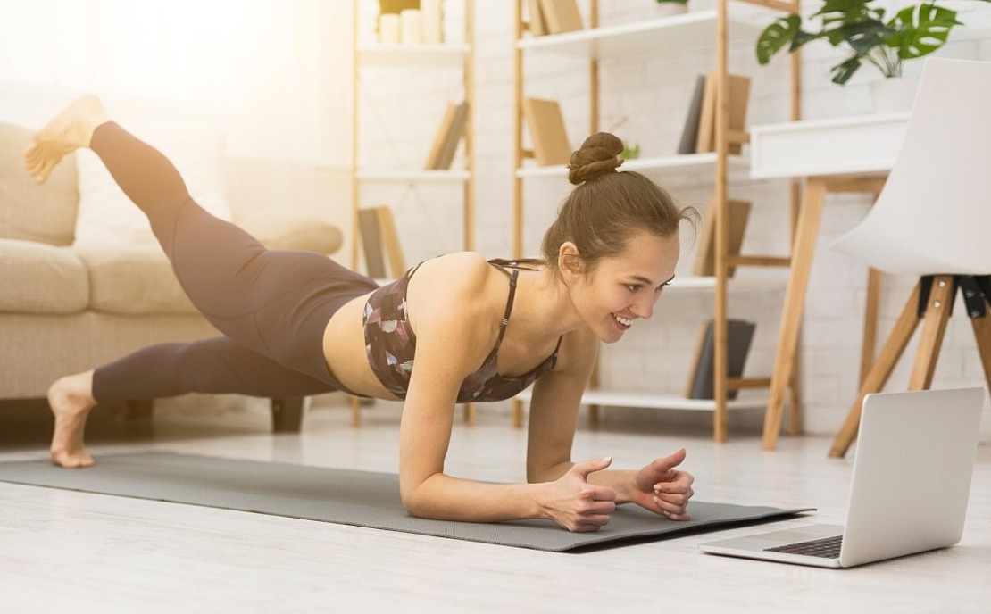 Eine Frau, die auf einer Yogamatte in ihrem Wohnzimmer eine Planke mit einem angehobenen Bein macht, während sie lächelt und auf den Laptop vor ihr schaut.