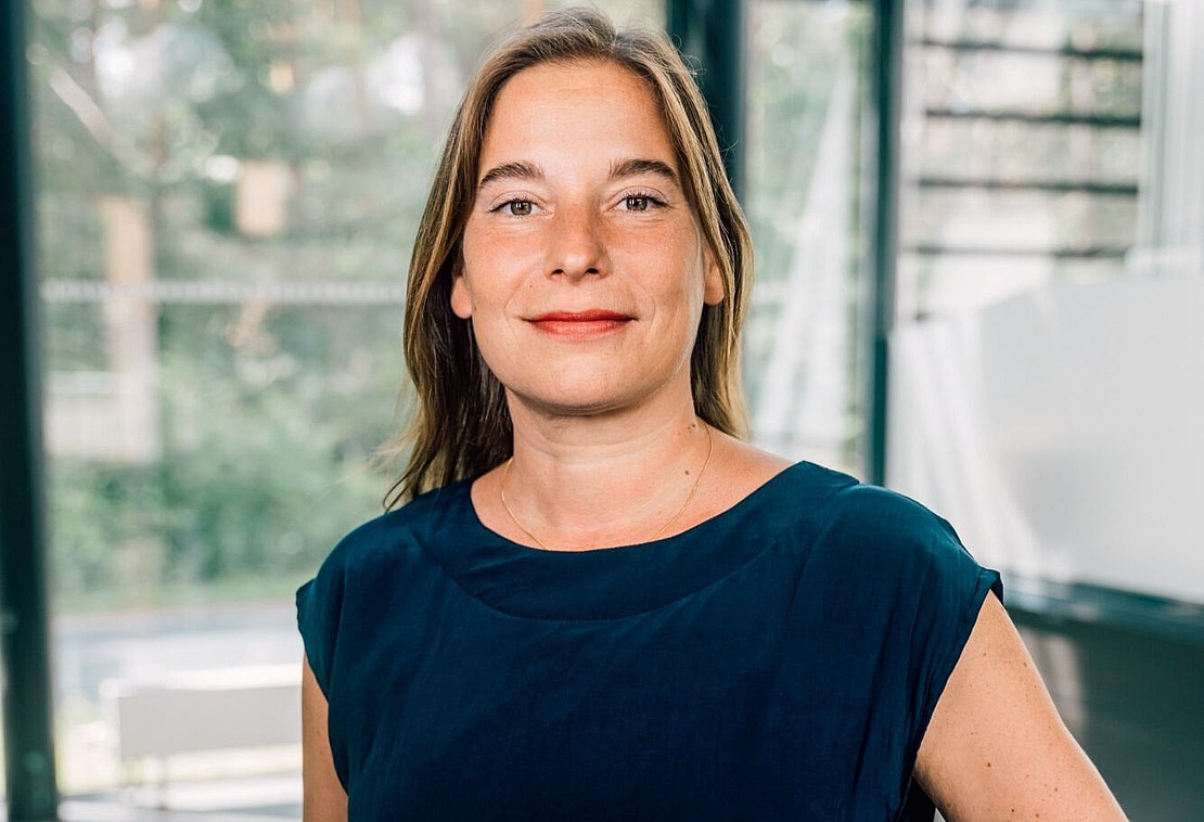 Eine Frau mit glattem, hellbraunem Haar steht in einem Haus, trägt ein marineblaues Kleid und eine weiße Uhr. Sie lächelt leicht und stützt eine Hand auf die Hüfte. Im Hintergrund sind helle Fenster und Hängelampen zu sehen.