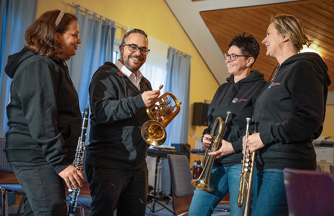 Vier Personen stehen in einem Raum, lächeln und halten Musikinstrumente, darunter eine Klarinette, eine Trompete und ein Saxophon. Sie scheinen sich in einem gut beleuchteten Raum mit hellblauen Vorhängen zu unterhalten.