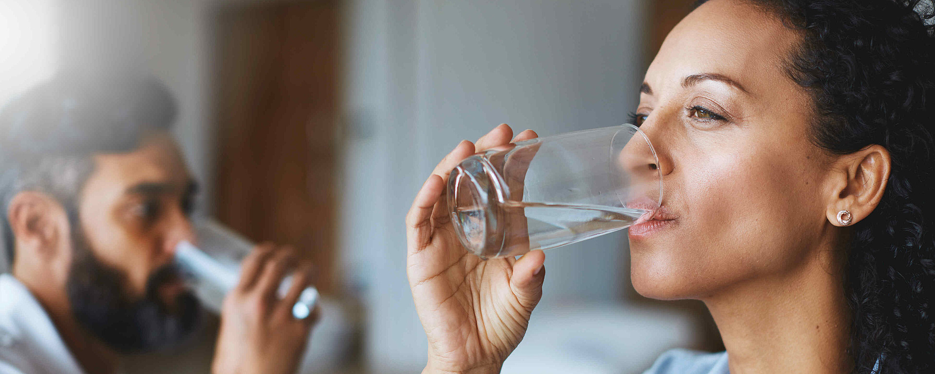 Eine Frau und ein Mann trinken jeweils ein Glas Wasser. Die Frau steht im Vordergrund, während der Mann unscharf im Hintergrund zu sehen ist. Beide wirken ruhig und auf das Trinken konzentriert.