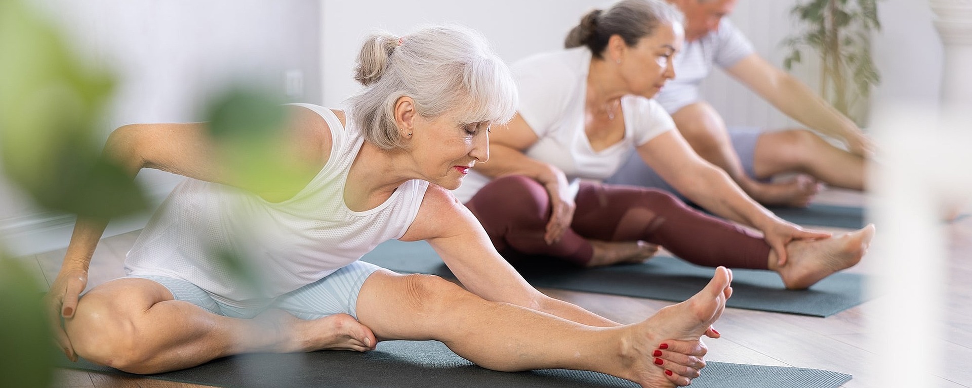 Drei ältere Erwachsene sitzen in einem Raum auf Yogamatten und strecken sich nach vorne, um ihre Zehen zu berühren. Sie tragen bequeme Trainingskleidung und wirken konzentriert und entspannt in einem hellen, luftigen Raum mit Holzboden.