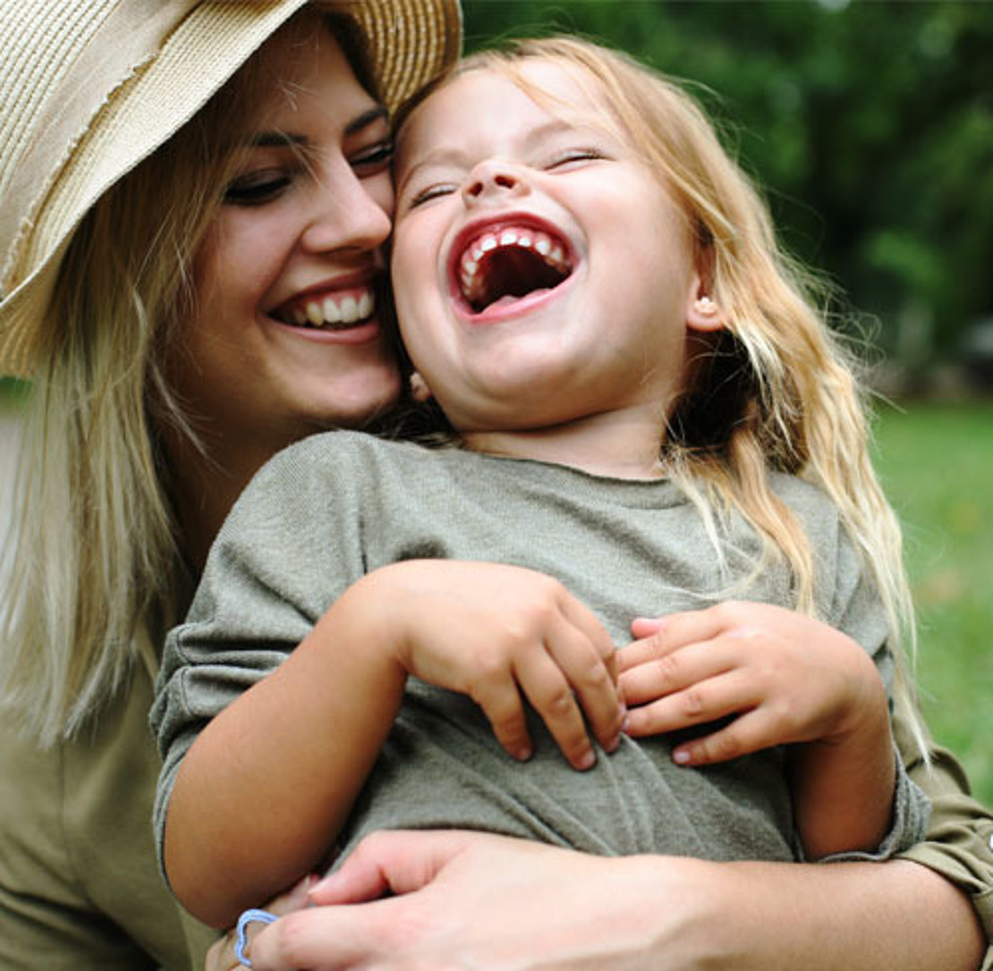 Eine Frau mit Hut lächelt und umarmt ein lachendes junges Mädchen im Freien, beide tragen legere grüne Kleidung. Der Hintergrund besteht aus unscharfem Grün, was vermuten lässt, dass sie sich in einem Park oder Garten befinden.