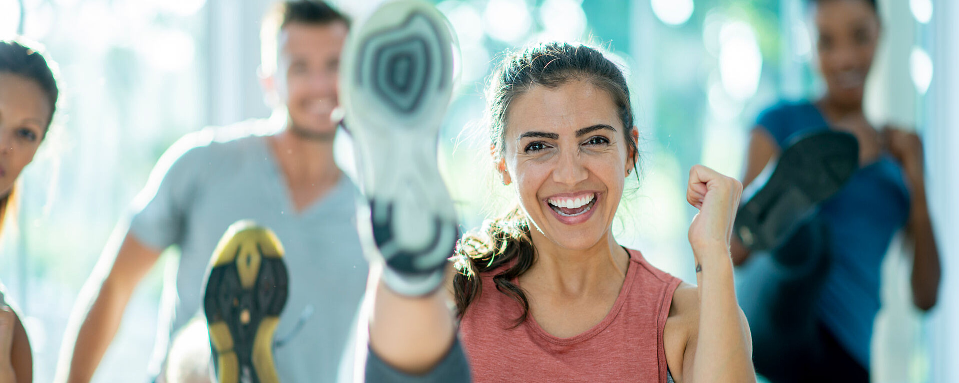 Eine Gruppe von Menschen treibt drinnen Sport, lächelt und schwingt die Beine hoch. Im Mittelpunkt steht eine Frau mit einem ärmellosen Oberteil, die energiegeladen und fröhlich aussieht, während die anderen im Hintergrund verschwommen sind.