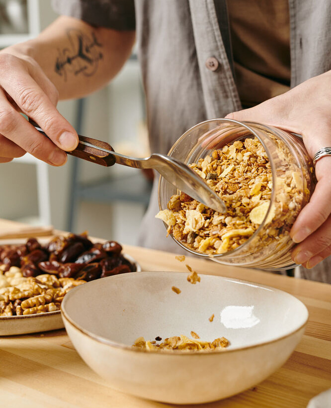 Eine Person schüttet mit einem Löffel Müsli aus einem Glasgefäß in eine weiße Schüssel. Ein Teller mit Datteln und Walnüssen steht daneben auf einer hölzernen Arbeitsplatte.