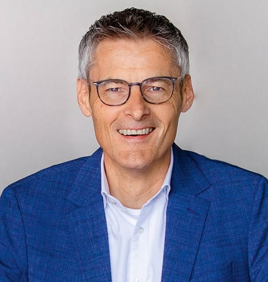 Ein Mann in einer blauen Anzugsjacke und mit Brille steht hinter einem geschwungenen, glänzenden Schreibtisch und lächelt in die Kamera vor einem hellgrauen Hintergrund mit zwei vertikalen Linien.