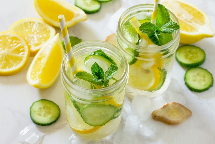 Zwei Einmachgläser, gefüllt mit Eiswasser, Zitronenscheiben, Gurkenscheiben und Minzblättern, jeweils mit einem gestreiften Strohhalm. Zitronenscheiben, Gurkenscheiben, Ingwer und Eiswürfel sind auf einer weißen Fläche verstreut.