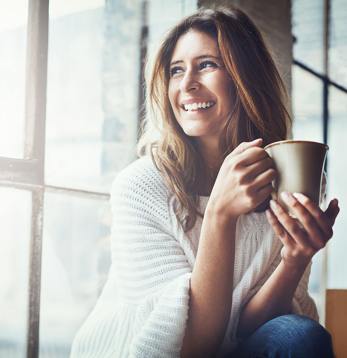 Eine Frau mit langem braunem Haar und einem weißen Pullover sitzt lächelnd an einem sonnenbeschienenen Fenster und hält mit beiden Händen eine Tasse.