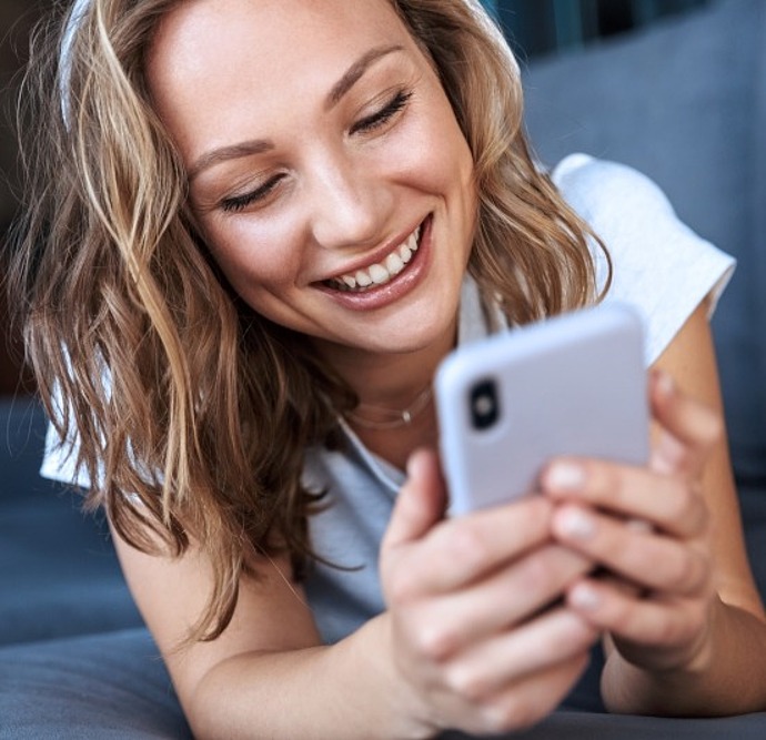 Eine junge Frau mit gewelltem blondem Haar lächelt, während sie auf einer Couch liegt und auf ihr Smartphone schaut. Sie wirkt entspannt und glücklich und hält das Telefon mit beiden Händen.