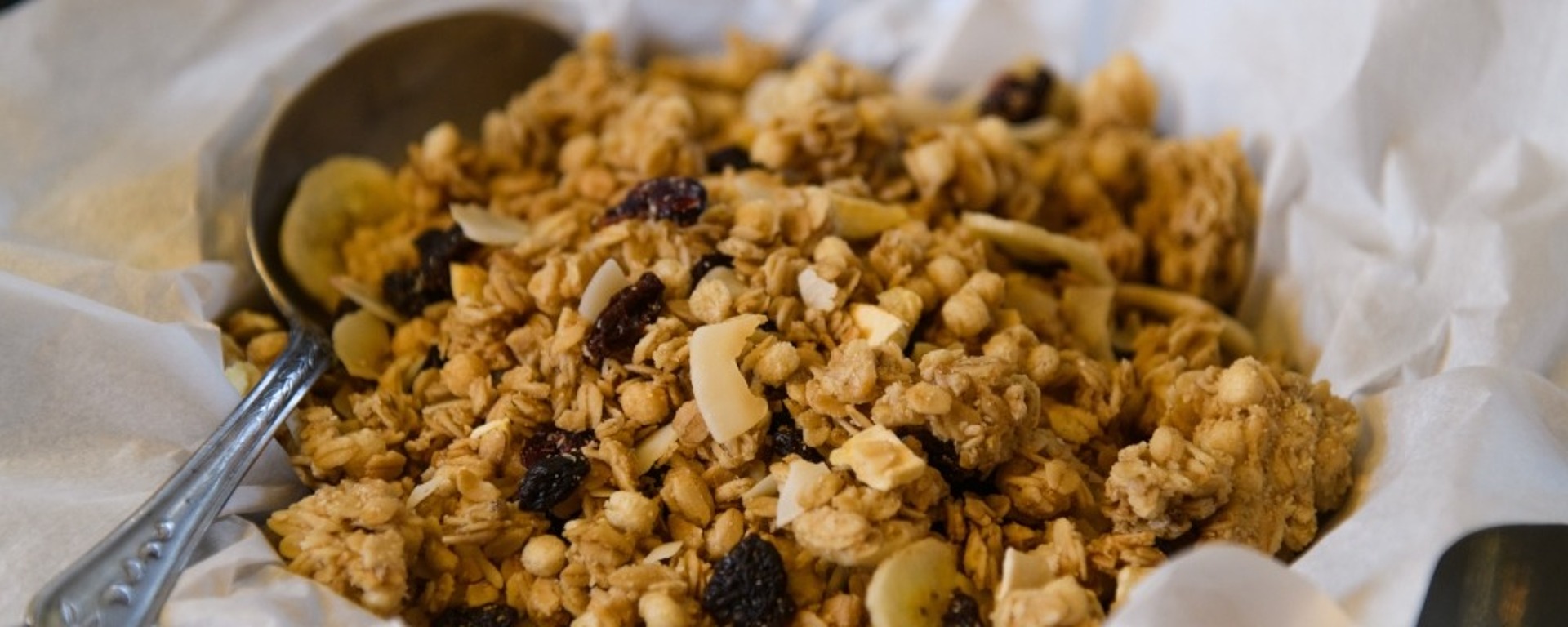 Eine Nahaufnahme einer Schüssel mit Müsli mit Hafer, Rosinen und Trockenfrüchten, die auf weißem Pergamentpapier steht. An der Seite sind teilweise zwei Metalllöffel zu sehen.