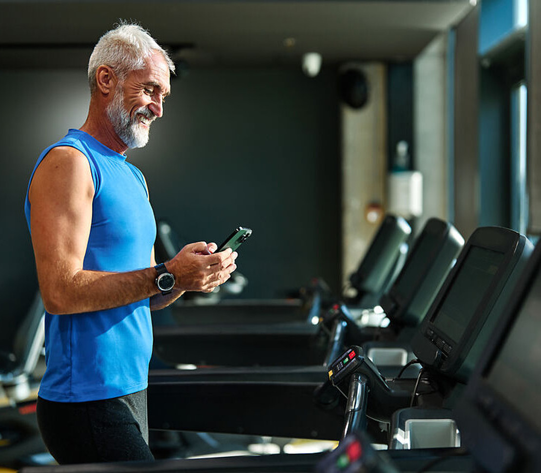 Ein älterer Mann in einem blauen, ärmellosen Shirt steht auf einem Laufband im Fitnessstudio und lächelt, während er auf sein Smartphone schaut. Im Hintergrund sind mehrere Laufbänder zu sehen.