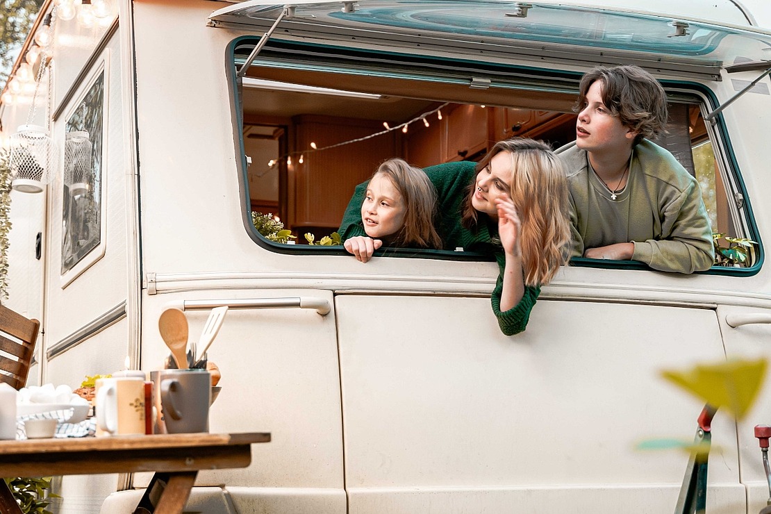 Drei Personen lehnen sich aus dem Fenster eines Wohnmobils, lächeln und schauen nach rechts. Im Vordergrund sind Küchenutensilien und ein Holztisch zu sehen. Die Atmosphäre ist gemütlich und entspannt, was an eine Camping- oder Road-Trip-Szene denken lässt.