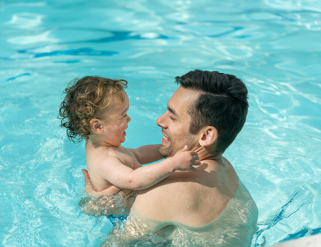 Ein Mann hält ein lächelndes Kleinkind in einem Schwimmbecken. Beide scheinen glücklich zu sein, wobei der Mann das Kind anschaut und das Kind sich an den Schultern des Mannes festhält. Das Wasser ist klar und blau.