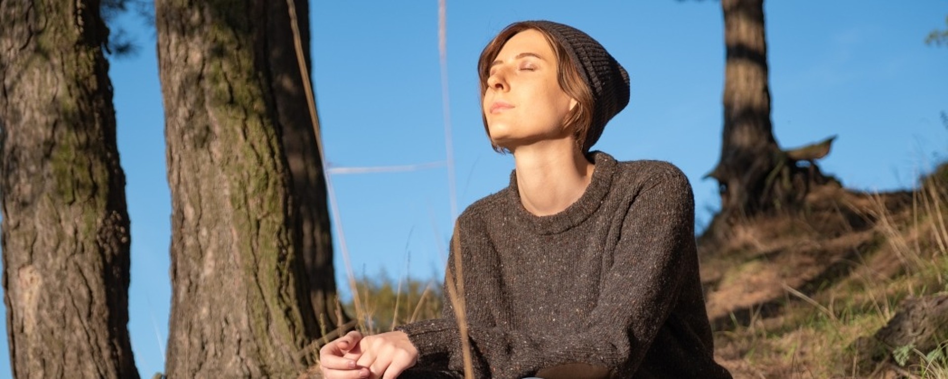 Eine Person, die einen dunklen Pullover, blaue Jeans und eine Strickmütze trägt, sitzt im Gras in der Nähe von Bäumen, einen Rucksack an der Seite, die Augen geschlossen und genießt das Sonnenlicht an einem klaren Tag.
