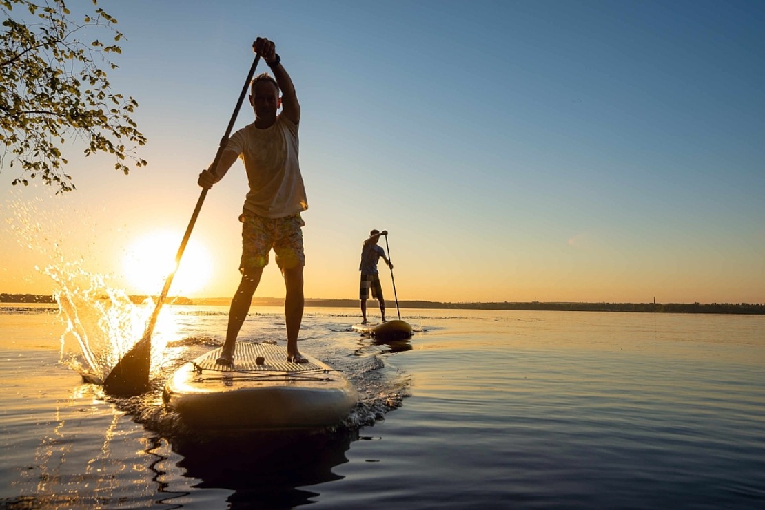 Zwei Paddelboarder auf einem ruhigen See bei Sonnenuntergang, mit Wasser, das von einem Paddel im Vordergrund plätschert, und der tiefstehenden Sonne am Horizont, die eine heitere und malerische Atmosphäre schafft.