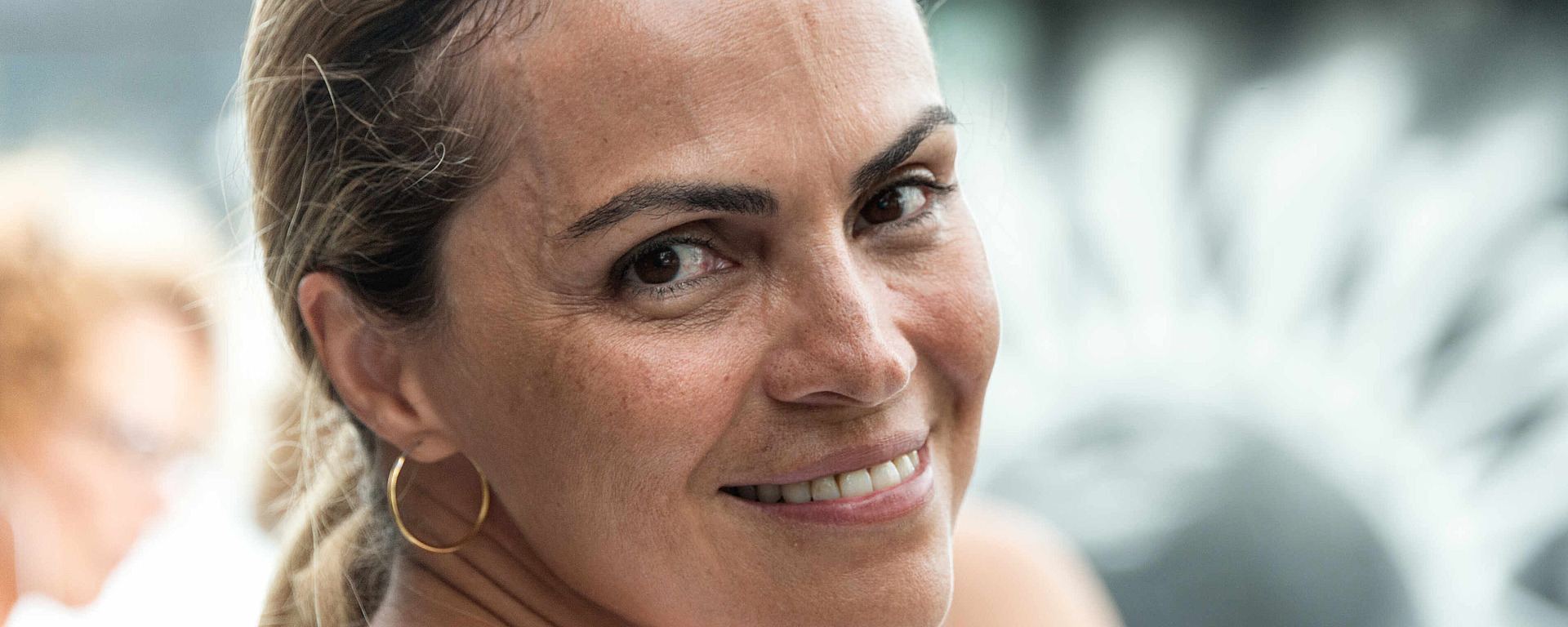 Eine Frau mit zurückgekämmtem hellbraunem Haar lächelt in die Kamera. Sie trägt ein weißes Oberteil und goldene Creolen. Der Hintergrund ist unscharf und zeigt eine weitere Person sowie ein kreisförmiges schwarz-weißes Muster.