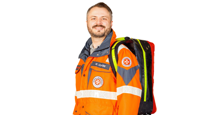 Ein Mann in einer orangefarbenen Notfalluniform mit reflektierenden Streifen und einem großen Rucksack lächelt vor einem schlichten weißen Hintergrund in die Kamera.