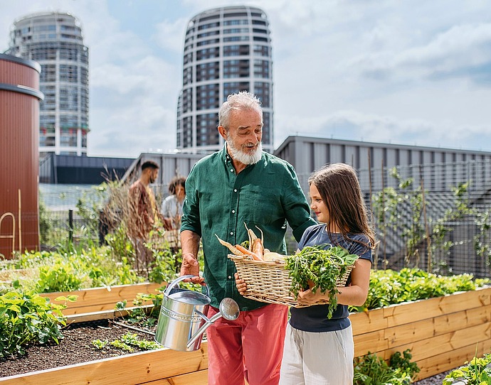 Ein älterer Mann und ein junges Mädchen stehen in einem städtischen Garten. Der Mann hält eine Gießkanne, während das Mädchen einen Korb mit frischem Gemüse trägt. Im Hintergrund erheben sich moderne Gebäude unter einem teilweise bewölkten Himmel.