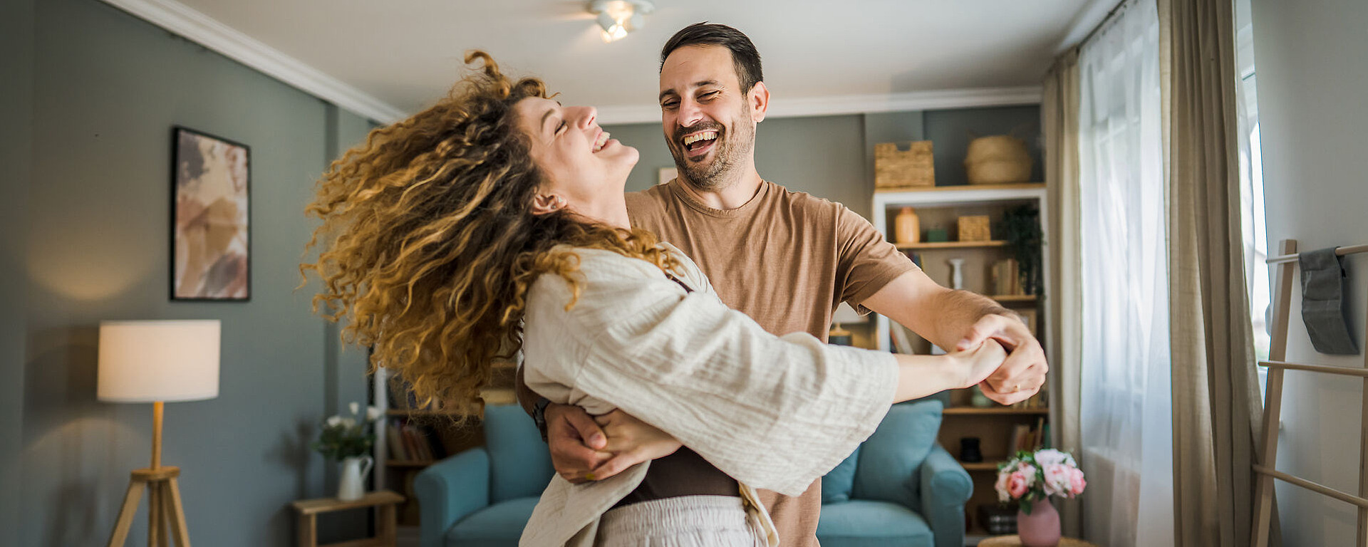 Ein Paar tanzt fröhlich zusammen in einem gemütlichen Wohnzimmer. Die Frau mit den lockigen Haaren lächelt breit, während der ebenfalls lächelnde Mann sie im Kreis dreht. Im Raum stehen ein blaugrünes Sofa, eine Lampe und dekorative Regale im Hintergrund.