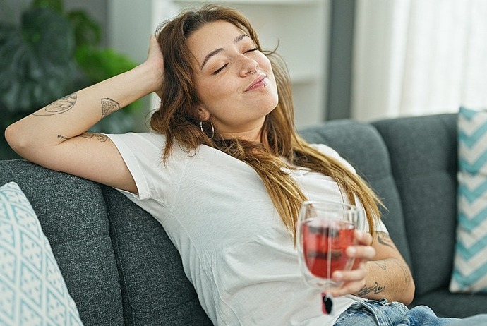 Eine junge Frau mit langen Haaren und Tattoos entspannt sich auf einer grauen Couch, lächelt mit geschlossenen Augen und hält ein Glas mit einem roten Getränk in der Hand. Sie sieht zufrieden aus und fühlt sich wohl in einer ungezwungenen häuslichen Umgebung.