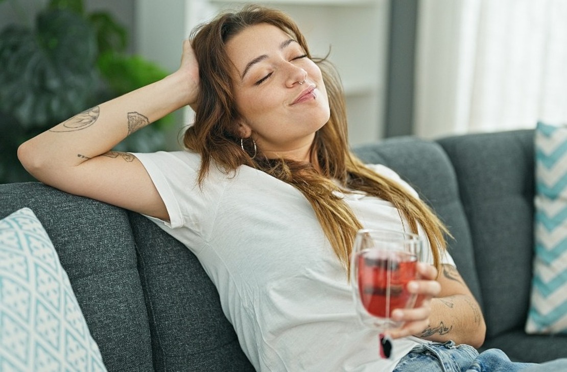 Eine junge Frau mit langen Haaren und Tattoos entspannt sich auf einer grauen Couch, lächelt mit geschlossenen Augen und hält ein Glas mit einem roten Getränk in der Hand. Sie sieht zufrieden aus und fühlt sich wohl in einer ungezwungenen häuslichen Umgebung.