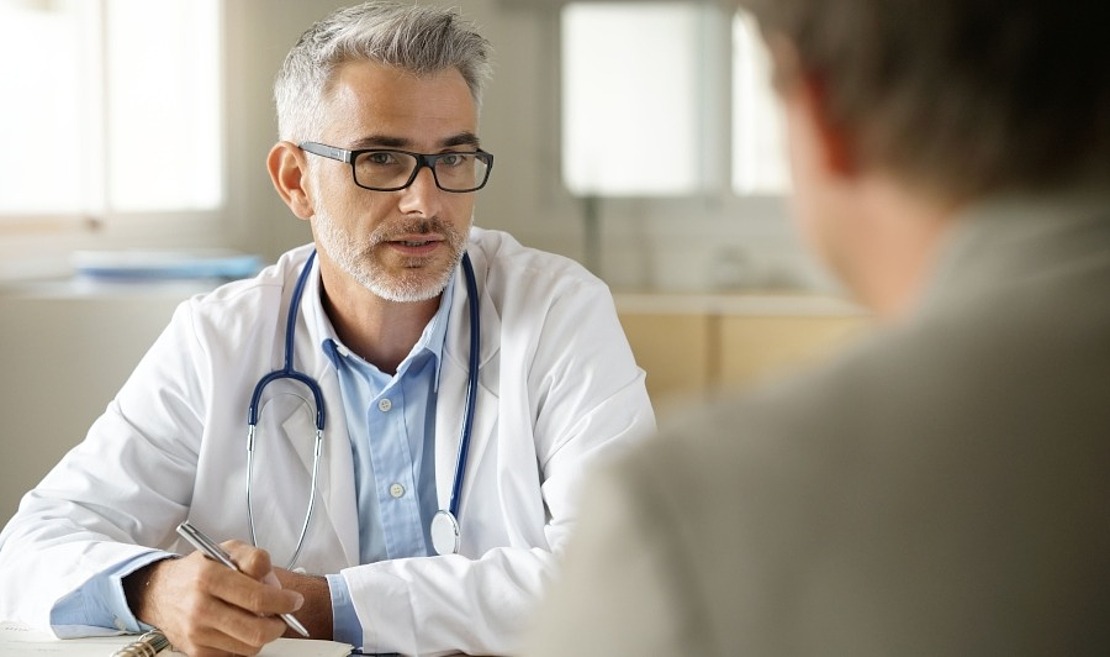 Ein männlicher Arzt mit grauem Haar und Brille, weißem Kittel und Stethoskop, spricht mit einem Patienten an einem Schreibtisch, der einen Stift und Notizen in der Hand hält.
