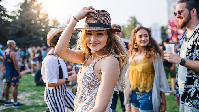 Eine junge Frau mit Hut und glitzerndem Tanktop lächelt bei einem Open-Air-Festival in die Kamera, umgeben von Menschen, die das Event an einem sonnigen Tag genießen.