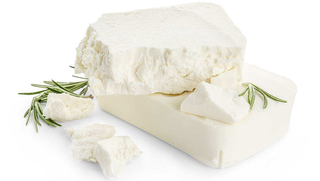 Zwei Blöcke weißer Fetakäse, einer davon teilweise zerbröselt, mit Rosmarinzweigen neben dem Käse und verstreuten Käsestücken auf weißem Hintergrund.