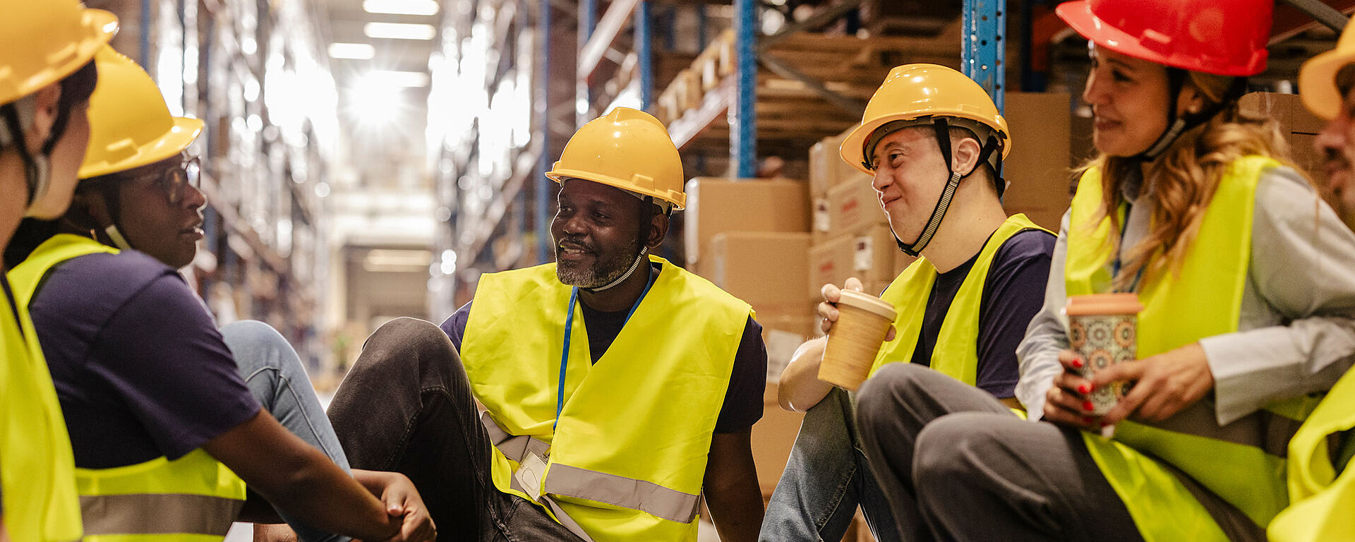 Eine Gruppe von Lagerarbeitern mit gelben Westen und Schutzhelmen sitzt während einer Pause zusammen auf dem Boden, lächelt und plaudert, während sie Kaffeetassen in einem mit Regalen und Kisten gefüllten Lagerbereich halten.