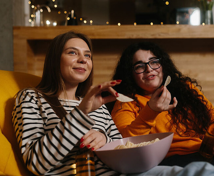 Zwei Frauen sitzen auf einer gelben Couch, essen Chips aus einer Schüssel und halten Getränke in der Hand, lächeln und beobachten etwas im Hintergrund. Der Hintergrund ist mit warmer Beleuchtung und Lichterketten versehen, die eine gemütliche Atmosphäre schaffen.