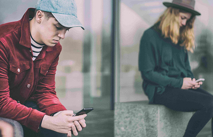 Eine Person in roter Jacke und Cap schaut auf ihr Handy, während sie neben einer Person mit Hut und grünem Pullover sitzt, die ebenfalls auf ihr Smartphone schaut. Sie sitzen auf einer Betonbank mit einer reflektierenden Glaswand im Hintergrund.