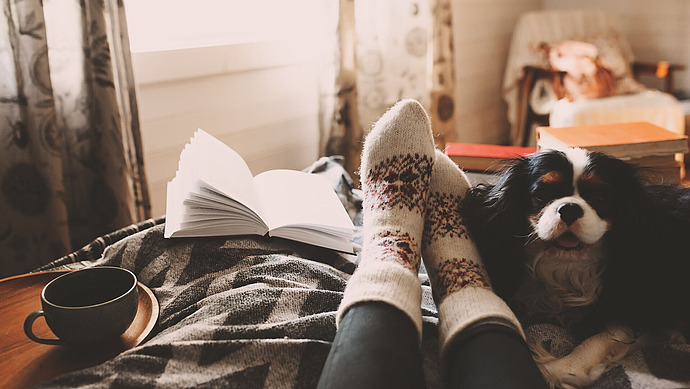 Eine Person mit gemusterten Socken entspannt sich auf einem gemütlichen Bett neben einem aufgeschlagenen Buch, einer Tasse Kaffee und einem Cavalier King Charles Spaniel, während sanftes Morgenlicht durch das Fenster fällt und warme Dekoration im Hintergrund zu sehen ist.