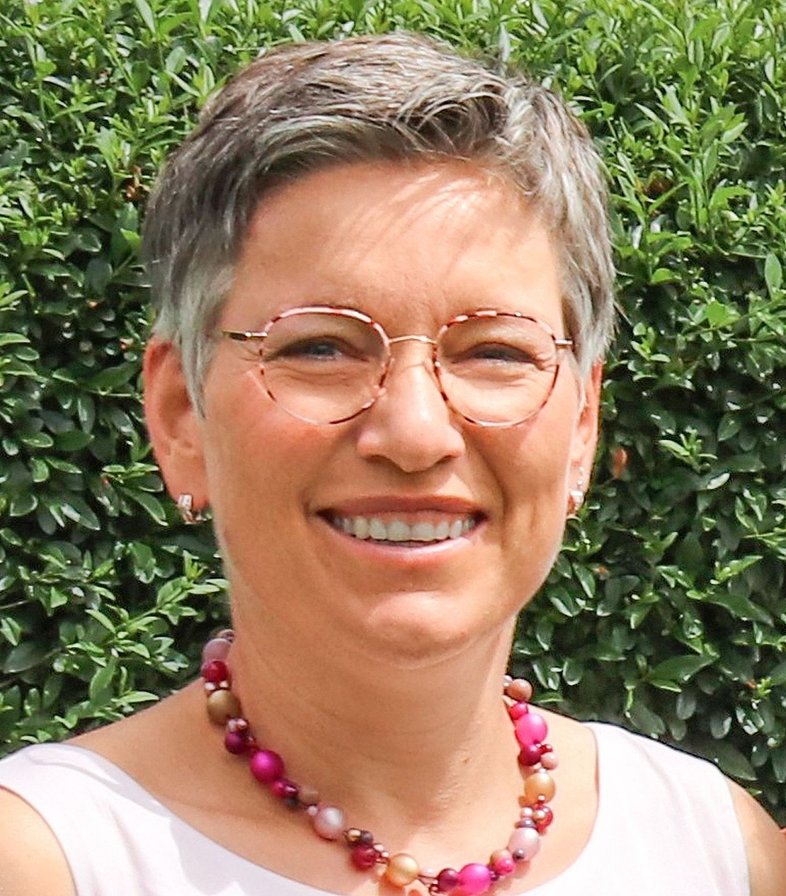 Eine lächelnde Person mit kurzen grauen Haaren, die eine runde Brille, eine rosafarbene Perlenkette und ein leichtes Oberteil trägt, steht im Freien vor einer grünen, belaubten Hecke.