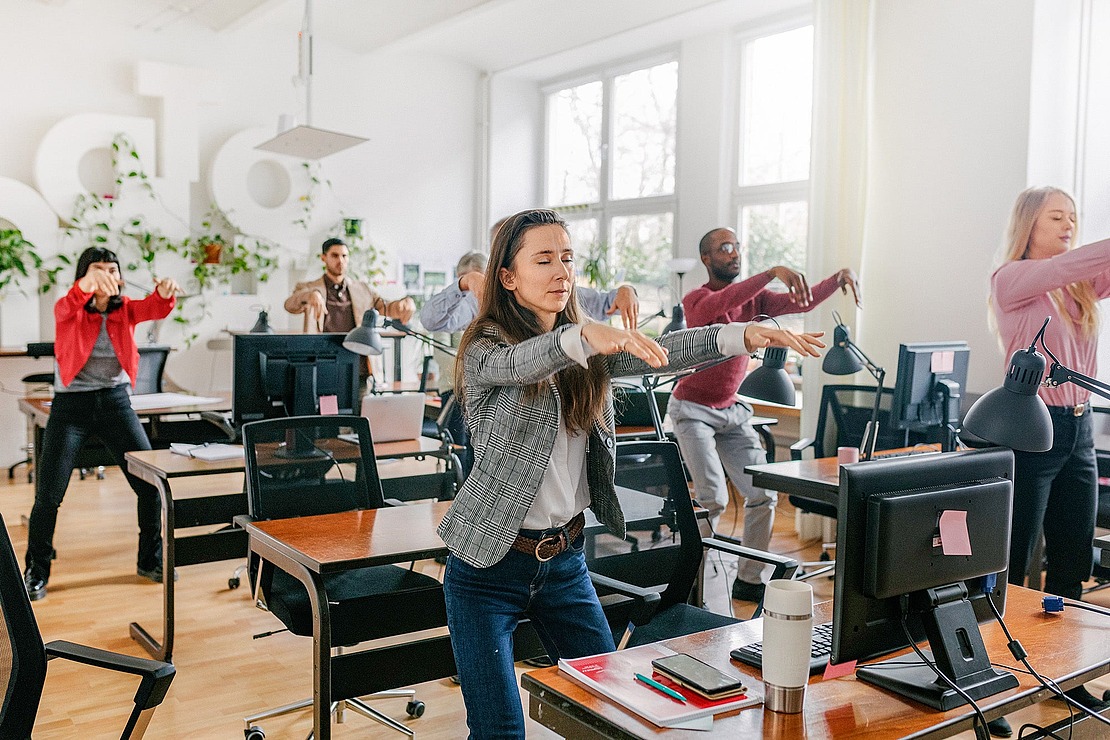 Eine Gruppe von Büroangestellten steht in der Nähe ihrer Schreibtische und macht gemeinsam Dehnungsübungen, wobei sie die Arme nach vorne streckt, in einem hellen, modernen Arbeitsraum mit Pflanzen und großen Fenstern.