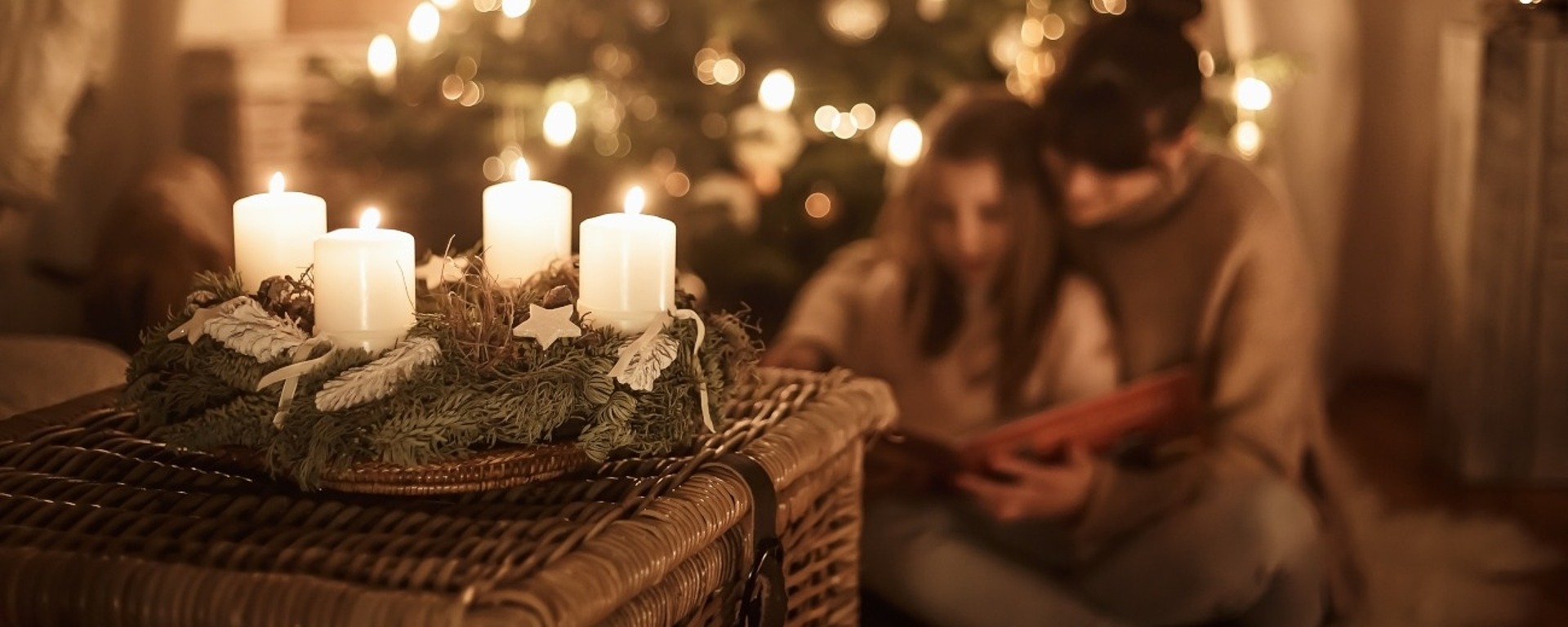 Ein gemütlicher, sanft beleuchteter Raum mit einem geschmückten Weihnachtsbaum im Hintergrund. Im Vordergrund stehen vier weiße Kerzen auf einem Kranz, der auf einer Weidentruhe steht. Zwei Personen, leicht verschwommen, sitzen lesend auf dem Boden zusammen.