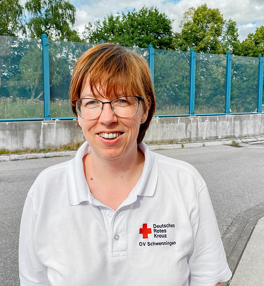 Eine lächelnde Person mit kurzen braunen Haaren und Brille steht im Freien auf einem gepflasterten Weg und trägt ein weißes Poloshirt mit dem Logo des Deutschen Roten Kreuzes. Im Hintergrund sind Bäume, ein blauer Himmel und eine Glaswand zu sehen.