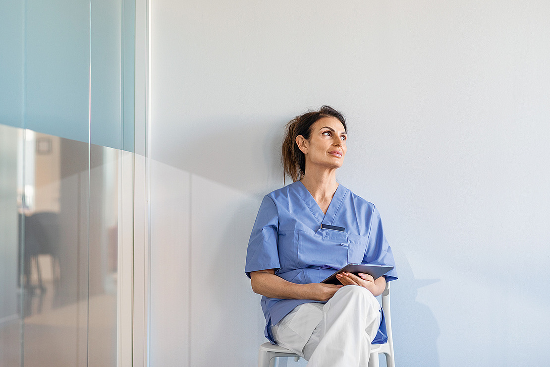 Eine Frau in blauem Arztkittel sitzt auf einem Stuhl vor einer weißen Wand, hält ein Tablet in der Hand und blickt nachdenklich nach oben.