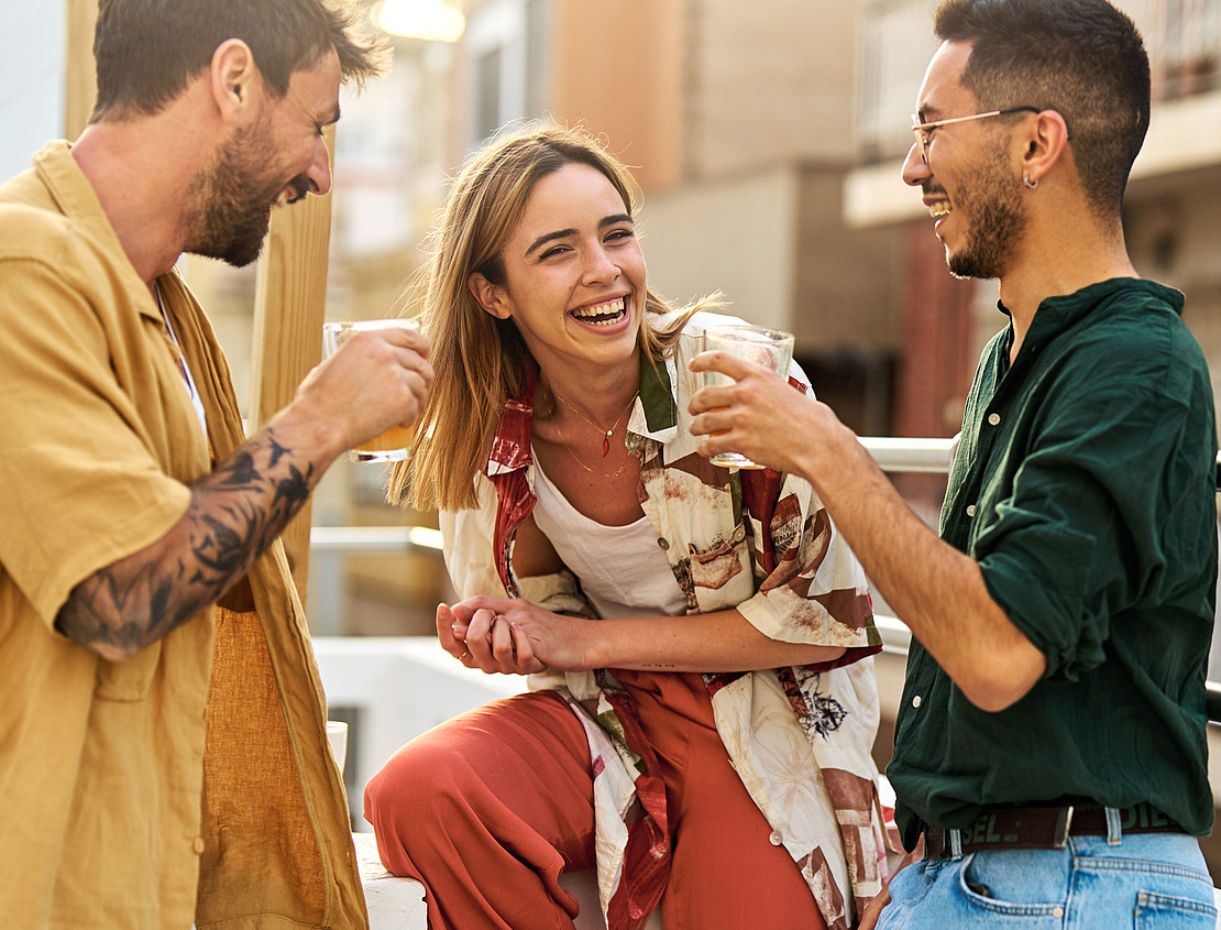 Drei junge Erwachsene lachen und genießen gemeinsam einen Drink auf einem sonnigen Balkon. In einer Sprechblase in der Ecke steht "MACH ES!" und ist angekreuzt.