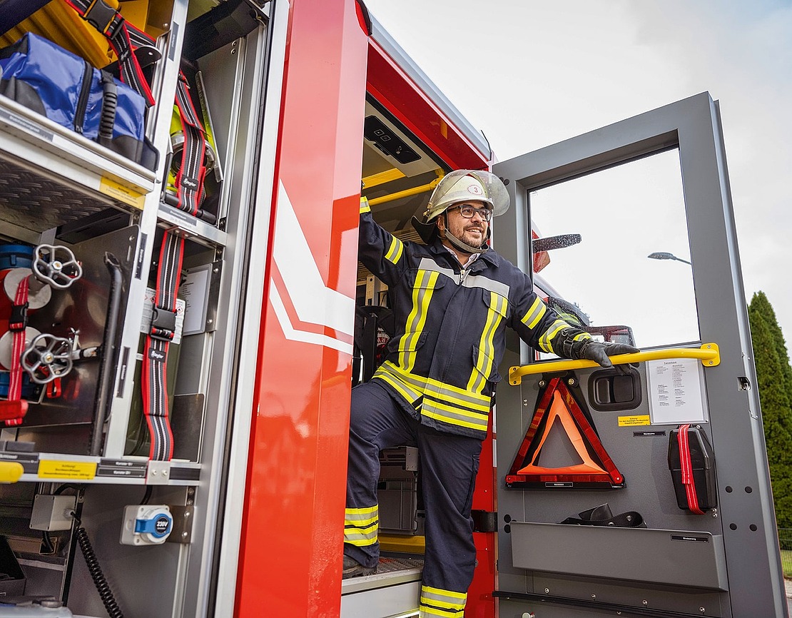 Ein Feuerwehrmann in voller Uniform und mit Helm steht in der offenen Tür eines Feuerwehrautos, lächelt und blickt nach außen, während im Inneren des Fahrzeugs die Feuerwehrausrüstung zu sehen ist.
