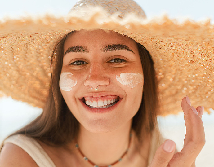 Eine lächelnde Frau mit großem Strohhut trägt am Strand Sonnencreme auf ihre Wangen auf. Sie hat langes braunes Haar, einen Nasenring und blickt an einem sonnigen Tag direkt in die Kamera.