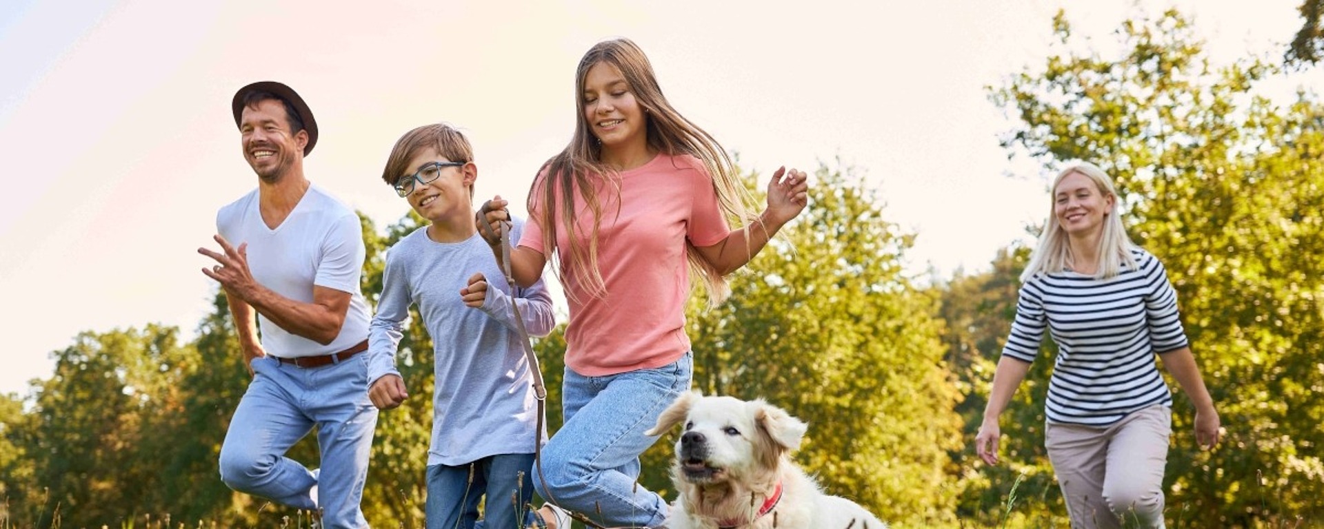 Eine Gruppe von vier Personen und ein Hund laufen an einem sonnigen Tag fröhlich über eine Wiese, die von grünen Bäumen umgeben ist. Alle sehen fröhlich und entspannt aus.