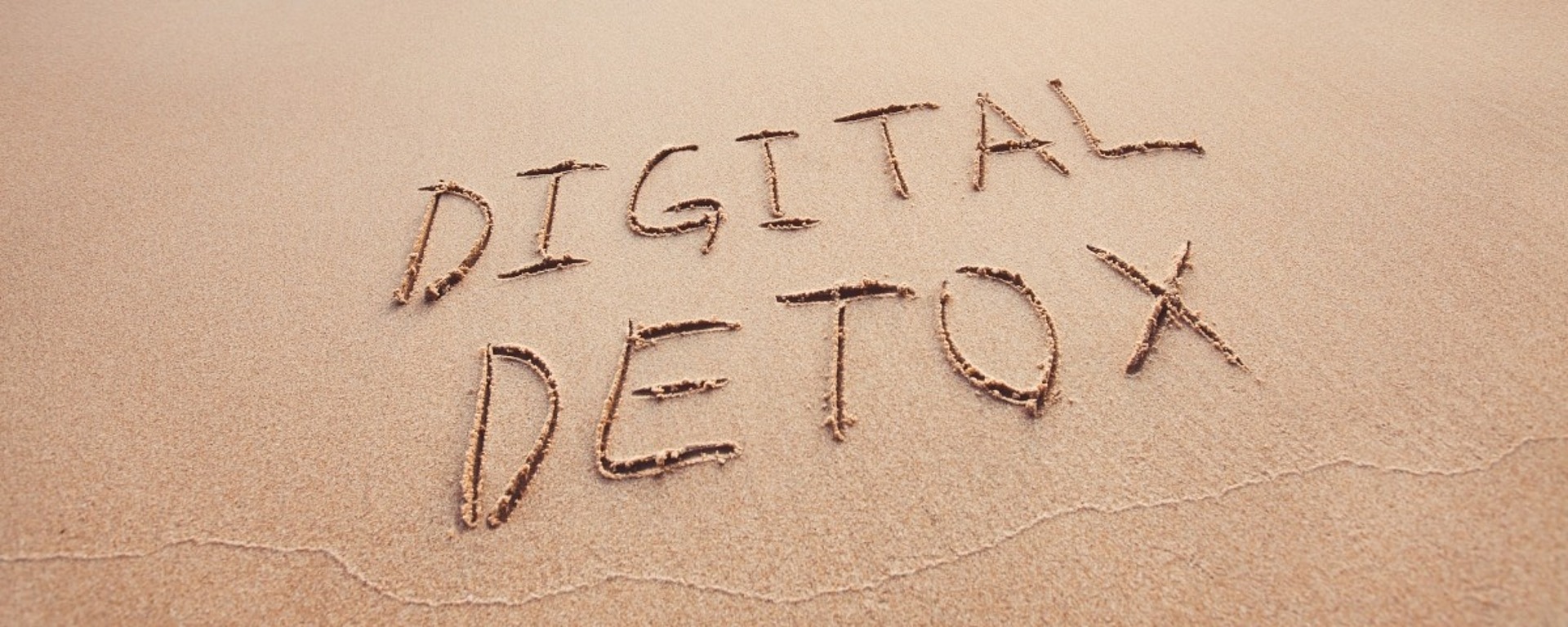 Die Worte "DIGITAL DETOX" sind in den Sand eines Strandes geschrieben, während sich im Hintergrund sanfte Meereswellen dem Ufer nähern.