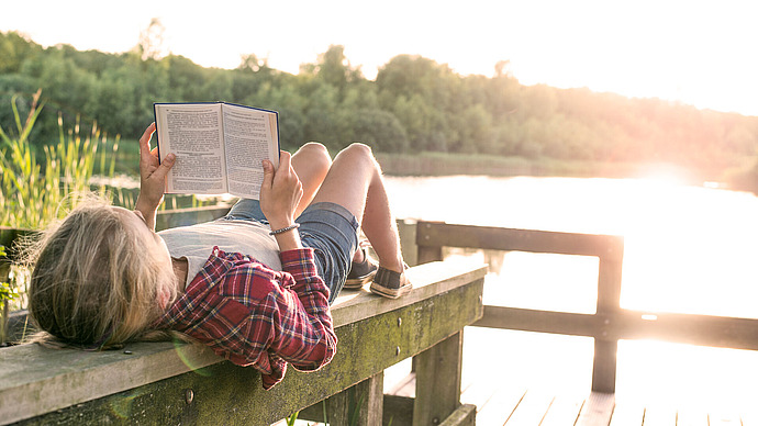 Eine Person liegt auf einem Holzsteg an einem See und liest ein Buch. Sie trägt ein kariertes Hemd und Shorts, die Sonne scheint hell über das Wasser und im Hintergrund sind grüne Bäume zu sehen.