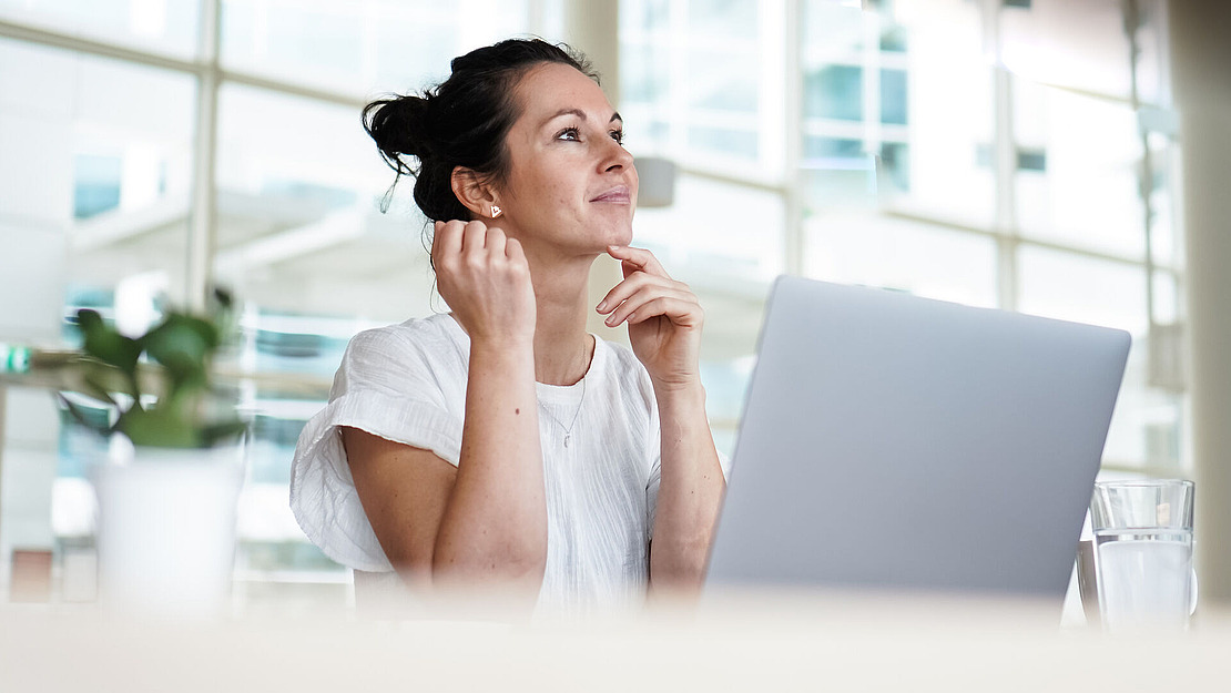 Eine Frau sitzt an einem Schreibtisch mit einem Laptop, lächelt und schaut nachdenklich auf. Sie trägt ein weißes Hemd, und in der Nähe stehen eine Topfpflanze und ein Glas Wasser. Im Hintergrund sind helle Fenster zu sehen.